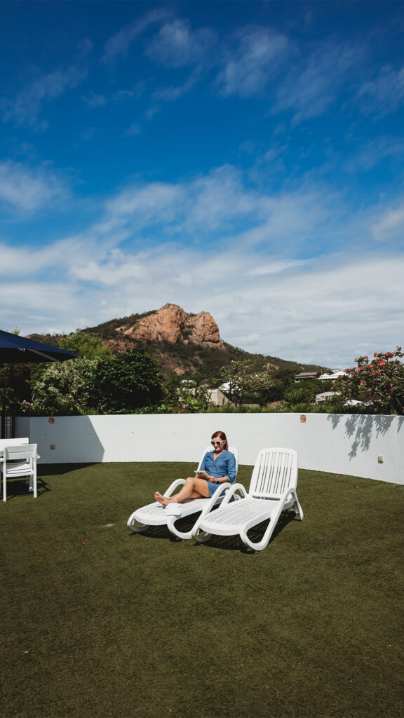A young woman sits on a lounge chair in her yard on a blue sky, sunny day with Castle Hill in the background.